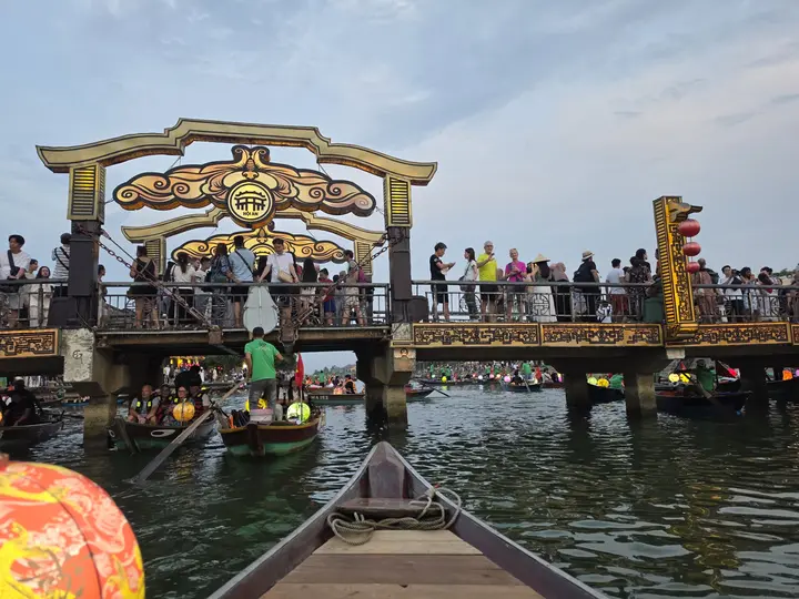 View from a wooden boat toward a decorated bridge crowded with people in Hoi An